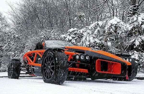 A black with orange highlights Ariel Atom parked in the snow. Behind it are snow-covered leafless trees.